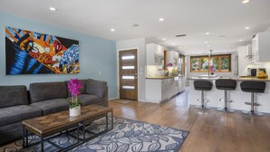 A modern living room with a sofa, coffee table, and a vibrant abstract painting. A kitchen with white cabinets and a bar area is visible in the background.