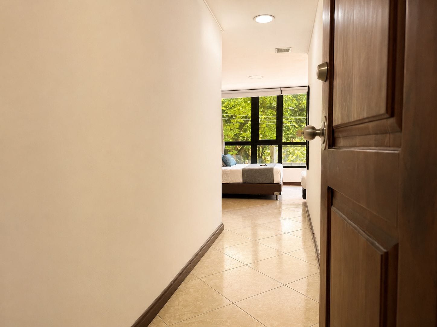 A hallway leading to a bedroom with a large window, a bed, and a nightstand.