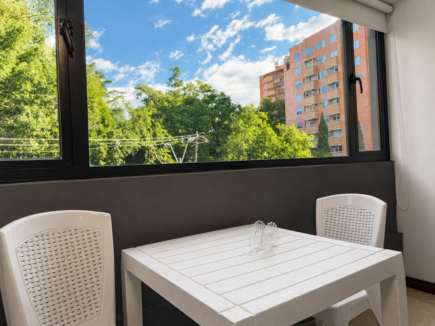 A balcony with a white table and two wicker chairs, overlooking a building and trees.