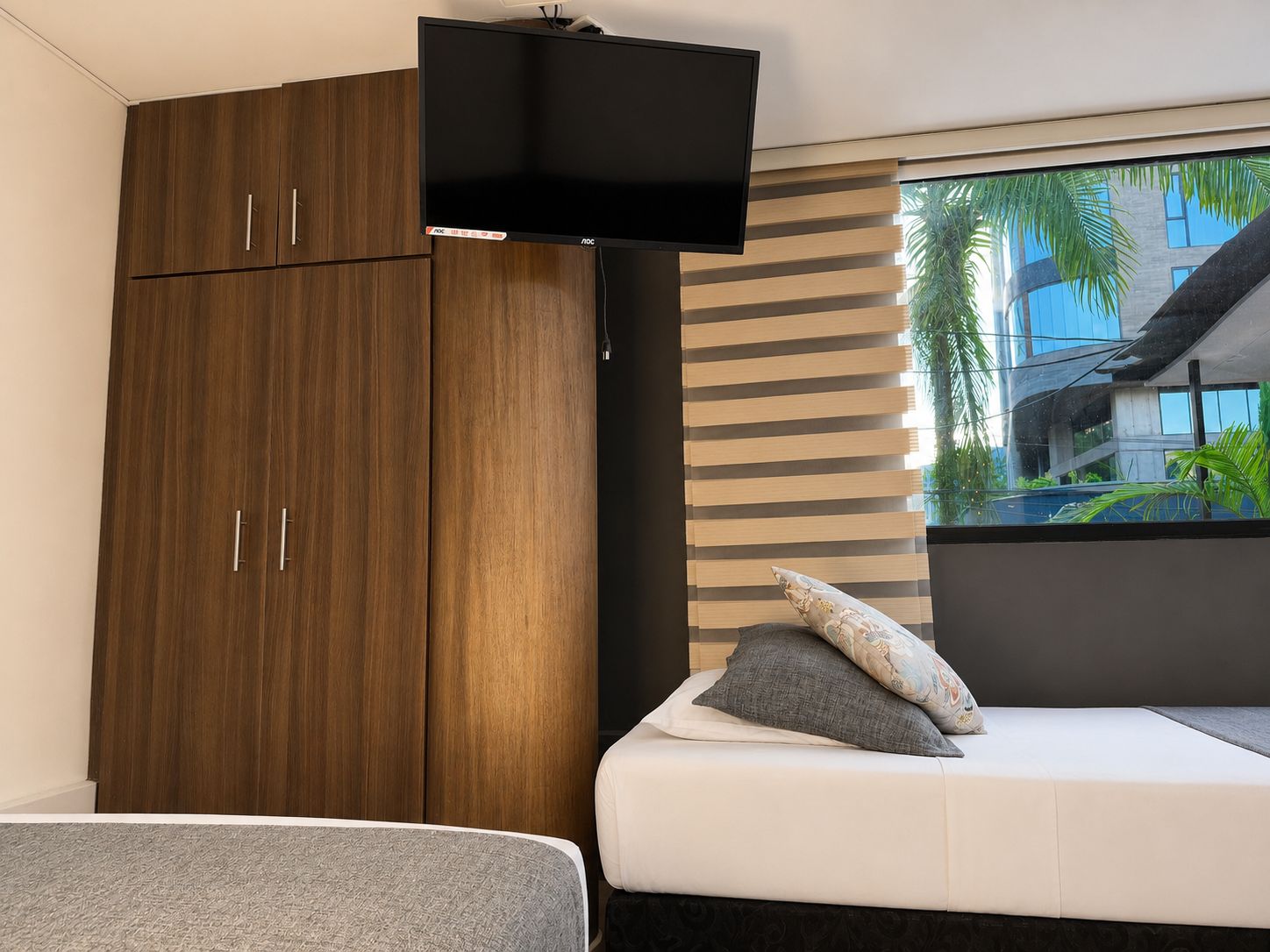 A bedroom with a bed, a TV mounted above it, and a window with blinds.
