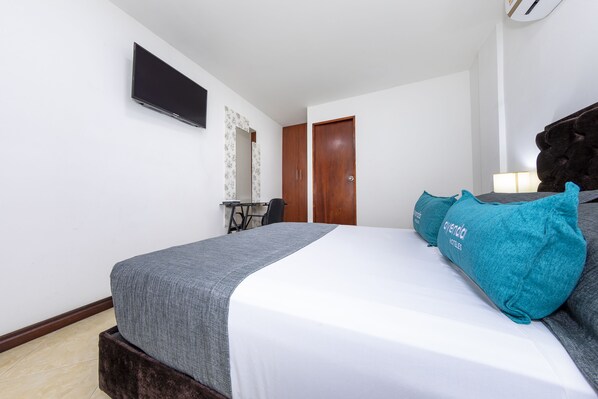 Een opgemaakt bed met een grijze deken en teal kussens, een flatscreen televisie aan de muur, een houten deur en een klein tafeltje met een stoel in de hoek.
