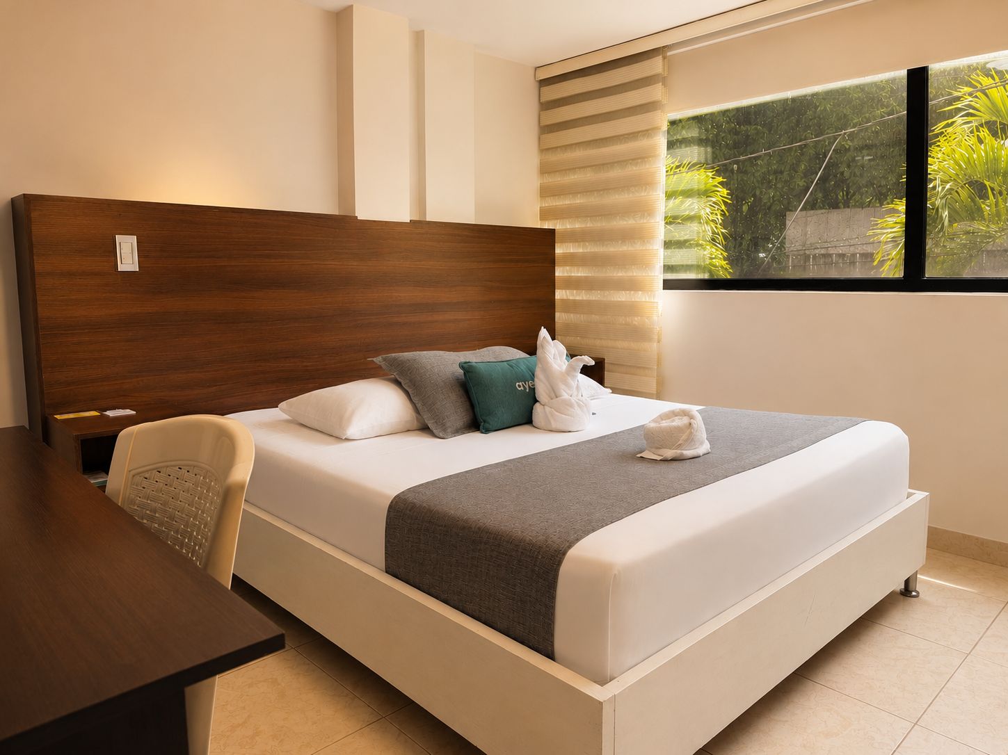 A hotel room with a wooden headboard, a bed with a grey blanket, two pillows, and a towel sculpture.