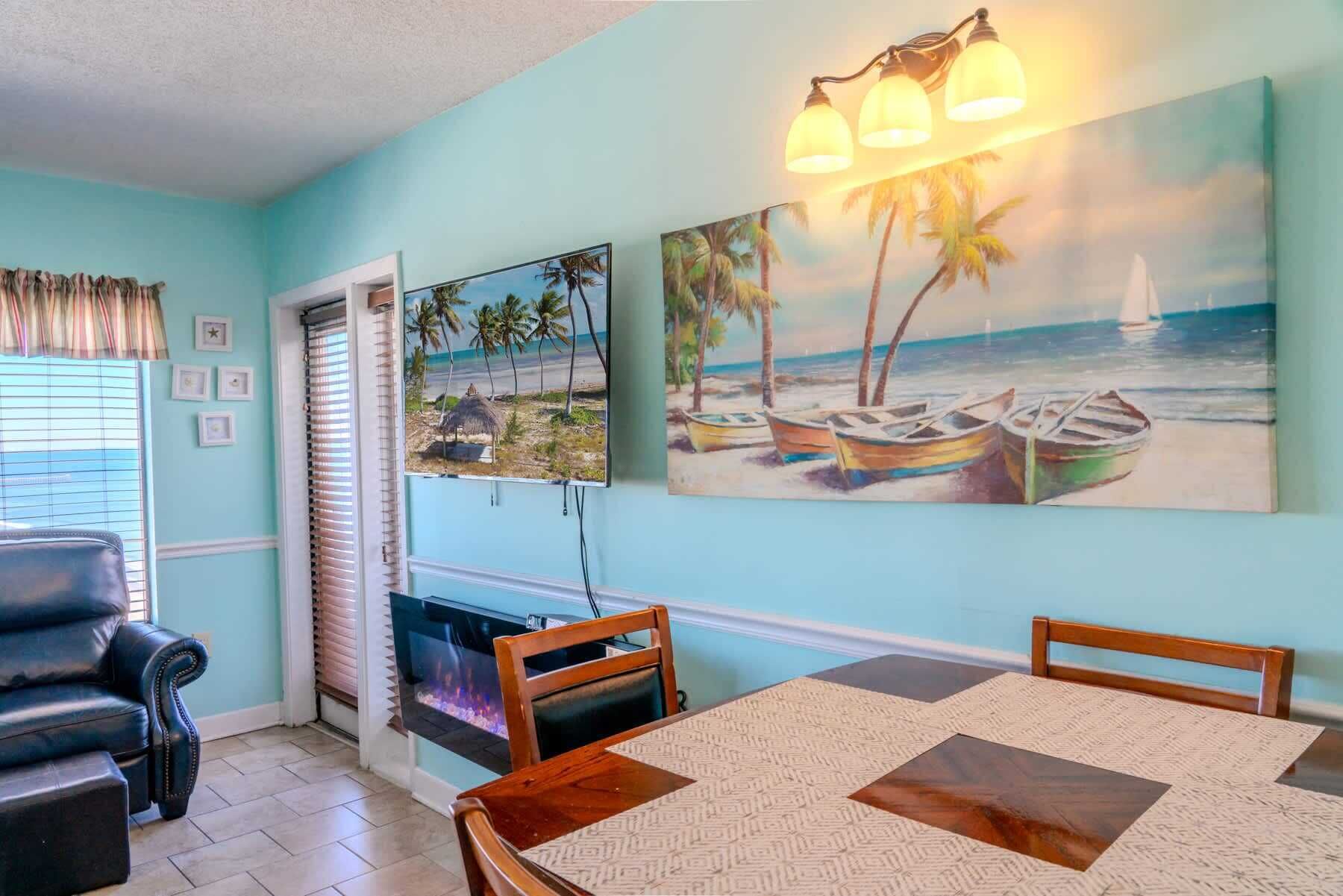 A dining area with a table, chairs, a fireplace, and two beach-themed paintings on the wall.