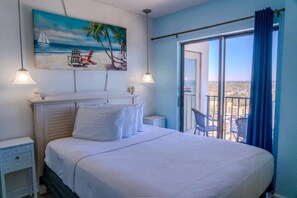 A bedroom with a bed, a painting of a beach scene, a balcony with chairs, and a view of the city.