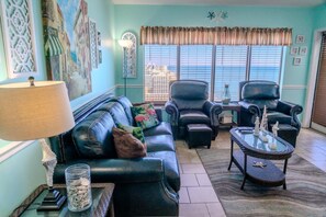 A living room with a sofa, two armchairs, a coffee table with model ships, a lamp, and a cityscape view through the windows.