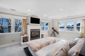 Un dormitorio con una cama grande, televisor, chimenea, telescopio y varias ventanas con vistas al agua.