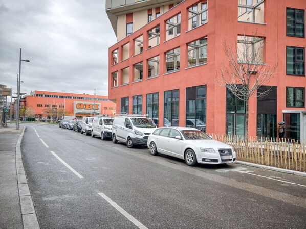 Ein modernes Gebäude mit roter Fassade und zahlreichen Fenstern. Vor dem Gebäude reihen sich mehrere weiße Autos an der Straße.