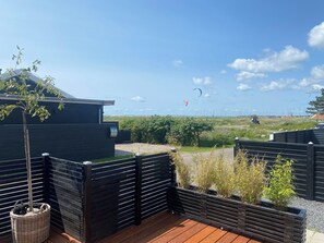 Eine Holzterrasse mit schwarzem Zaun, einem kleinen Baum und Blick auf den Himmel mit Drachen.