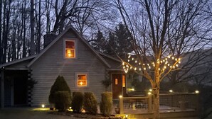 Una cabaña de troncos con dos ventanas iluminadas, un árbol decorado con luces y un camino de grava.