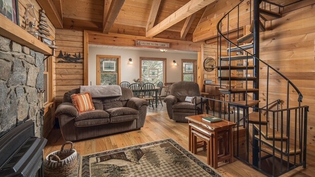 Interior acogedor de una cabaña con chimenea de piedra, piso de madera, un sofá, un sillón, una mesita y una escalera caracol.