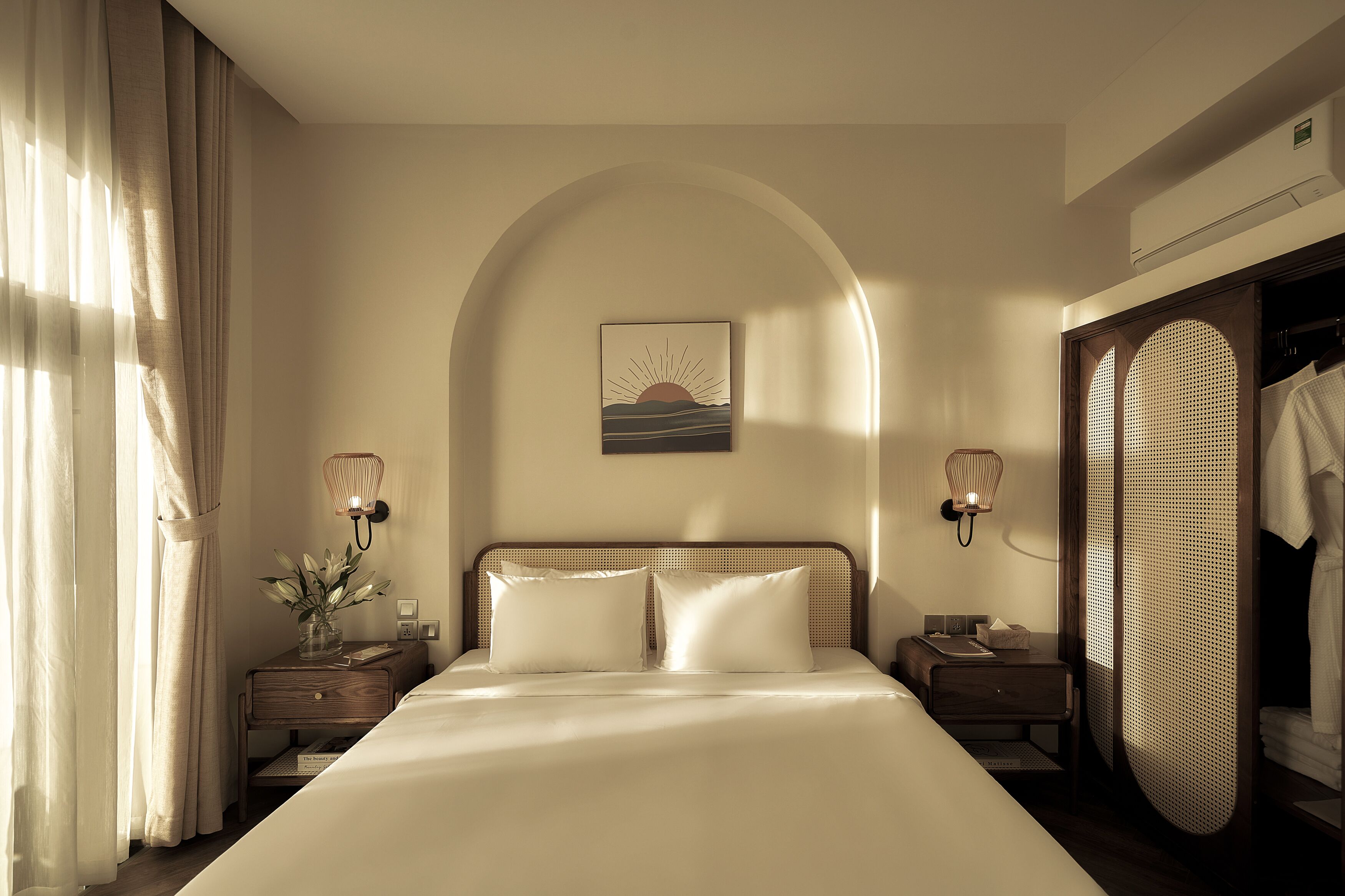A hotel room with a large bed, two bedside tables, a nightstand, a wall-mounted lamp, a decorative wall piece, and a wardrobe.