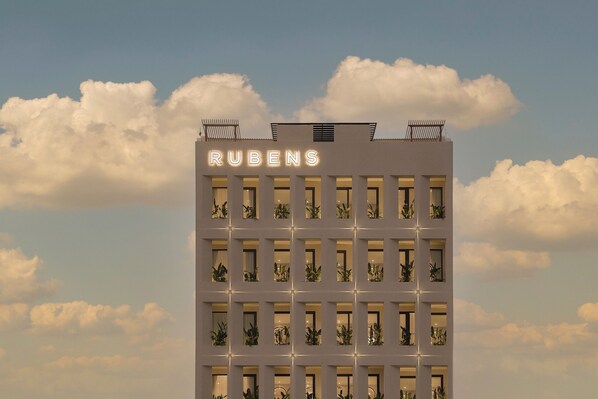 Een gebouw met de naam "RUBENS" verlicht op de gevel, met een raster van ramen en planten op de balkons.