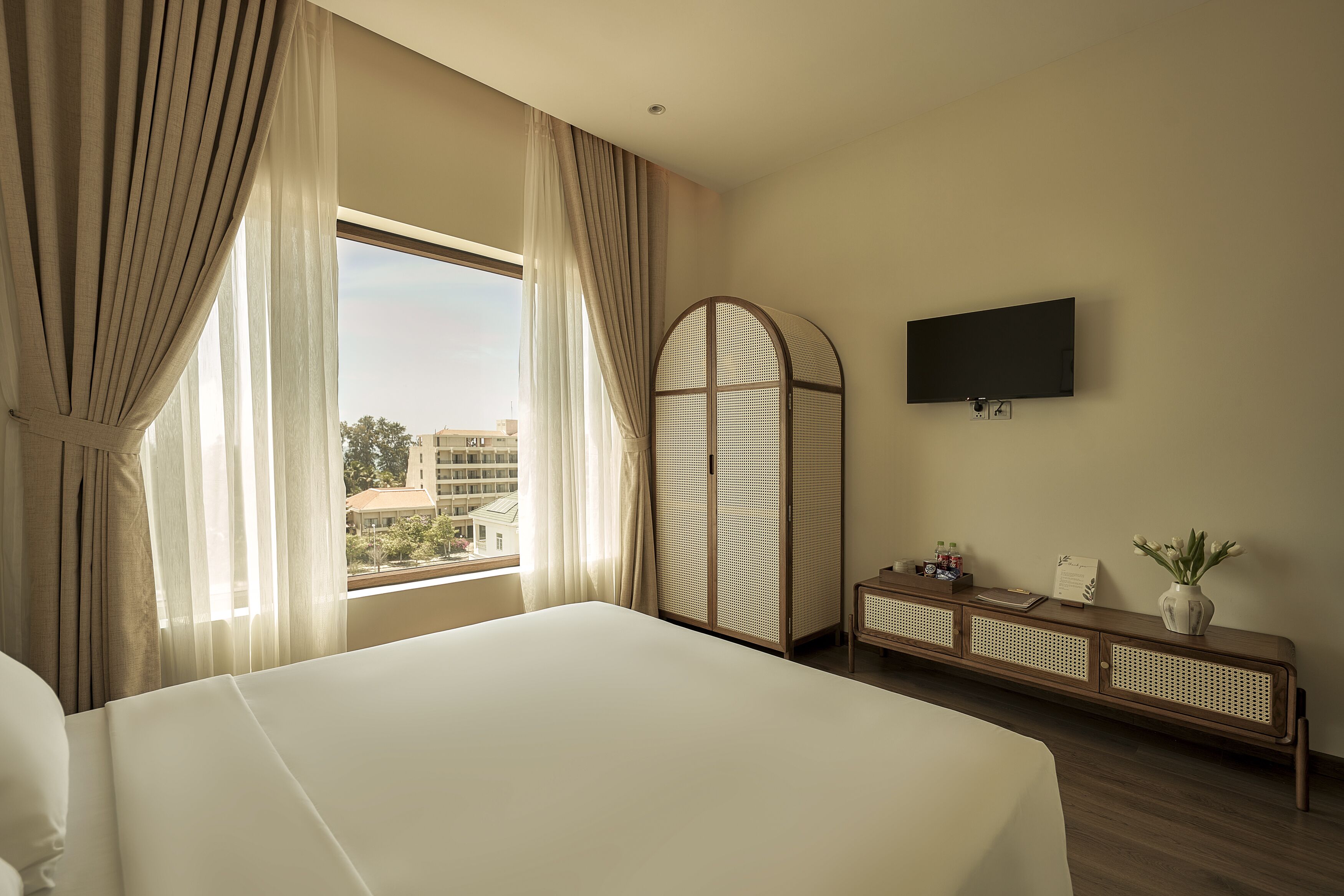 Ein Hotelzimmer mit einem großen Bett, einem an der Wand befestigten Fernseher, einem Holzkonsolentisch und Blick auf Gebäude vor dem Fenster.