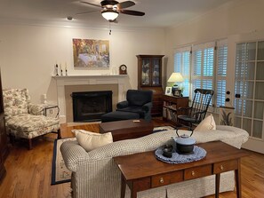 A living room with a fireplace, a ceiling fan, a painting, and a view through a window with blinds. - Martinez Masters Home (Martinez)