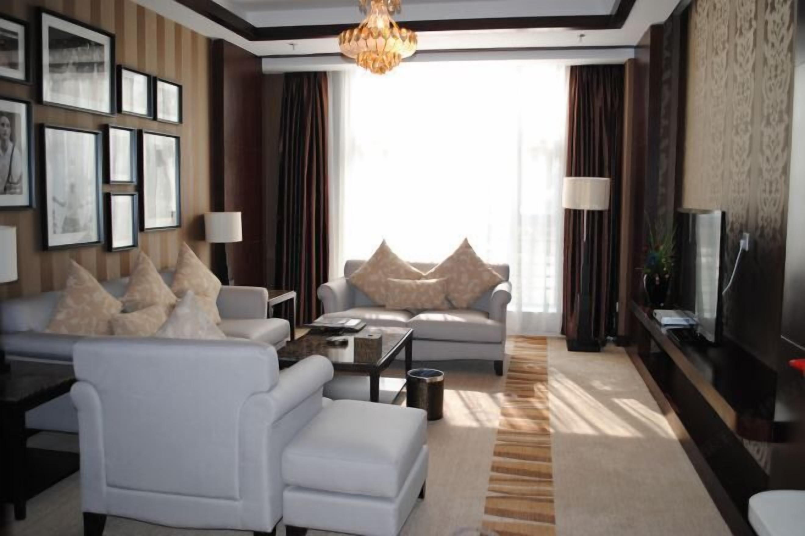 A modern living room with a sofa, armchair, coffee table, and a TV. There are framed pictures on the wall, a chandelier, and a large window with curtains.