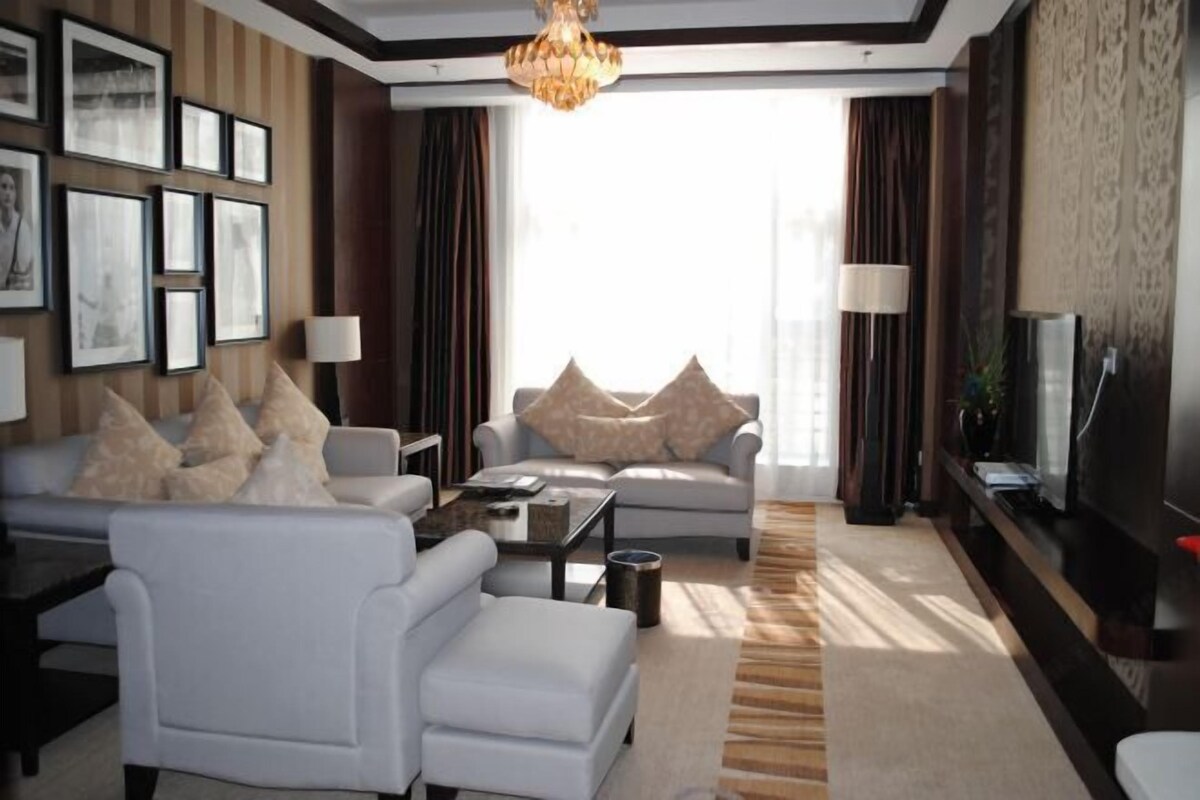 A modern living room with a sofa, armchair, coffee table, and a TV. There are framed pictures on the wall, a chandelier, and a large window with curtains.