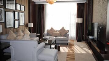A modern living room with a sofa, armchair, coffee table, and a TV. There are framed pictures on the wall, a chandelier, and a large window with curtains.