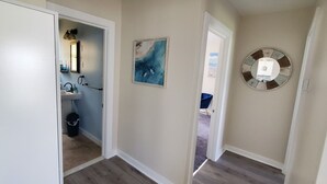 Un couloir avec une porte blanche, une salle de bain avec un lavabo et un miroir, et un mur orné d’un miroir rond et d’un tableau.