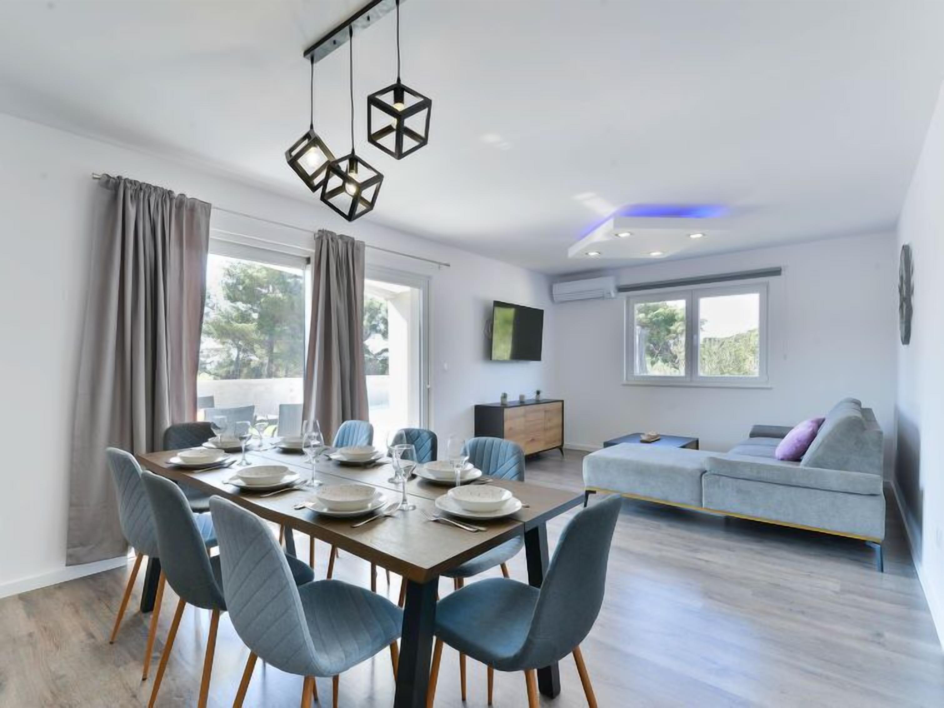 A modern dining area with a wooden table set for four, grey upholstered chairs, and a contemporary hanging light fixture.
