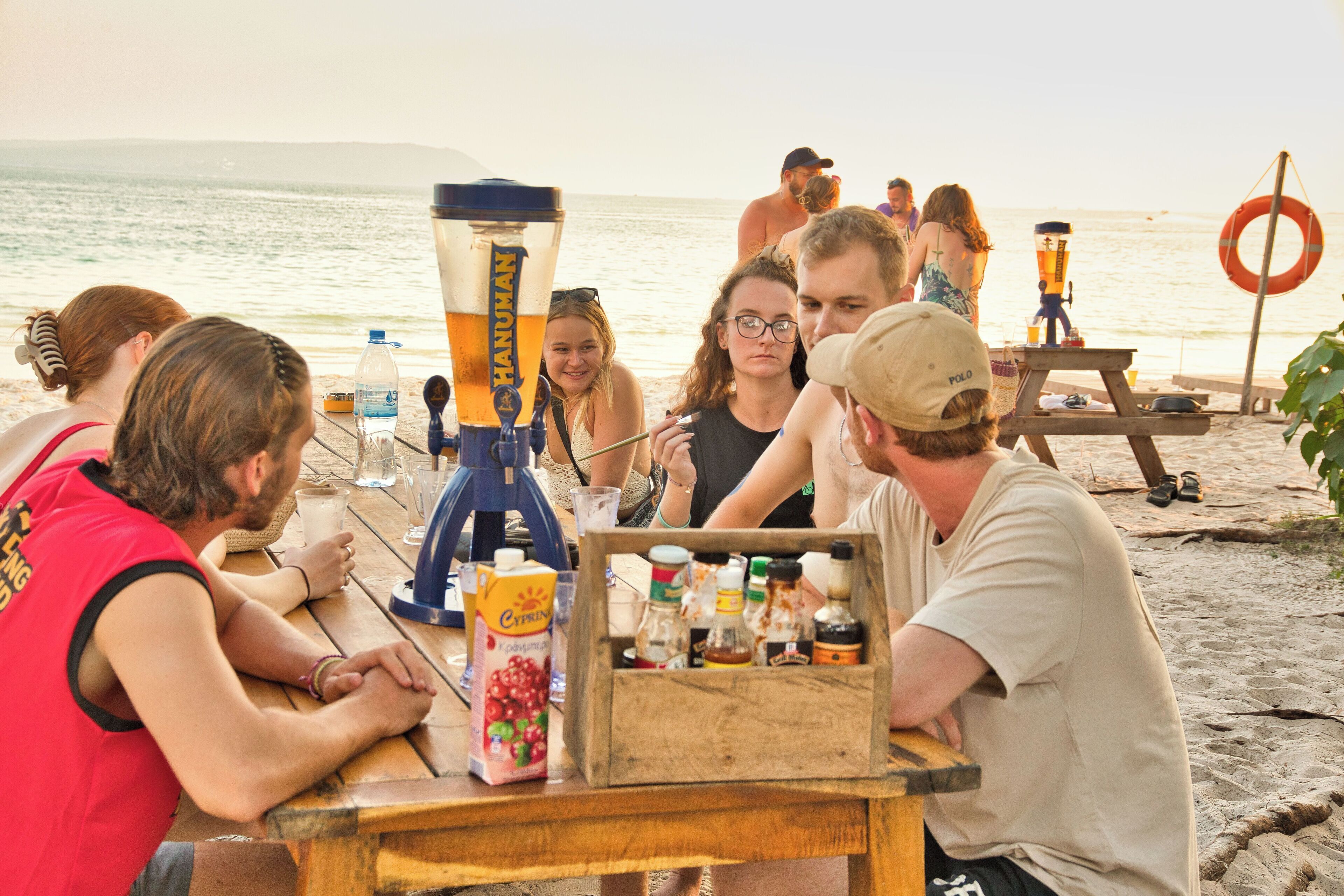 A group of people sitting at a wooden table on a beach, with a large beverage dispenser and a crate of bottles in the foreground.
