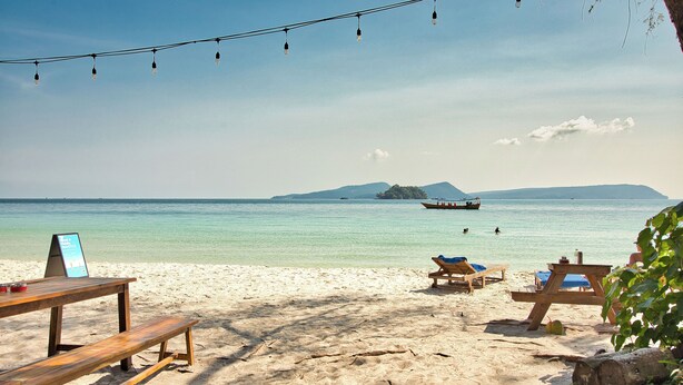 A beach with wooden benches and tables, a boat in the water, and a clear view of the sea and distant land.