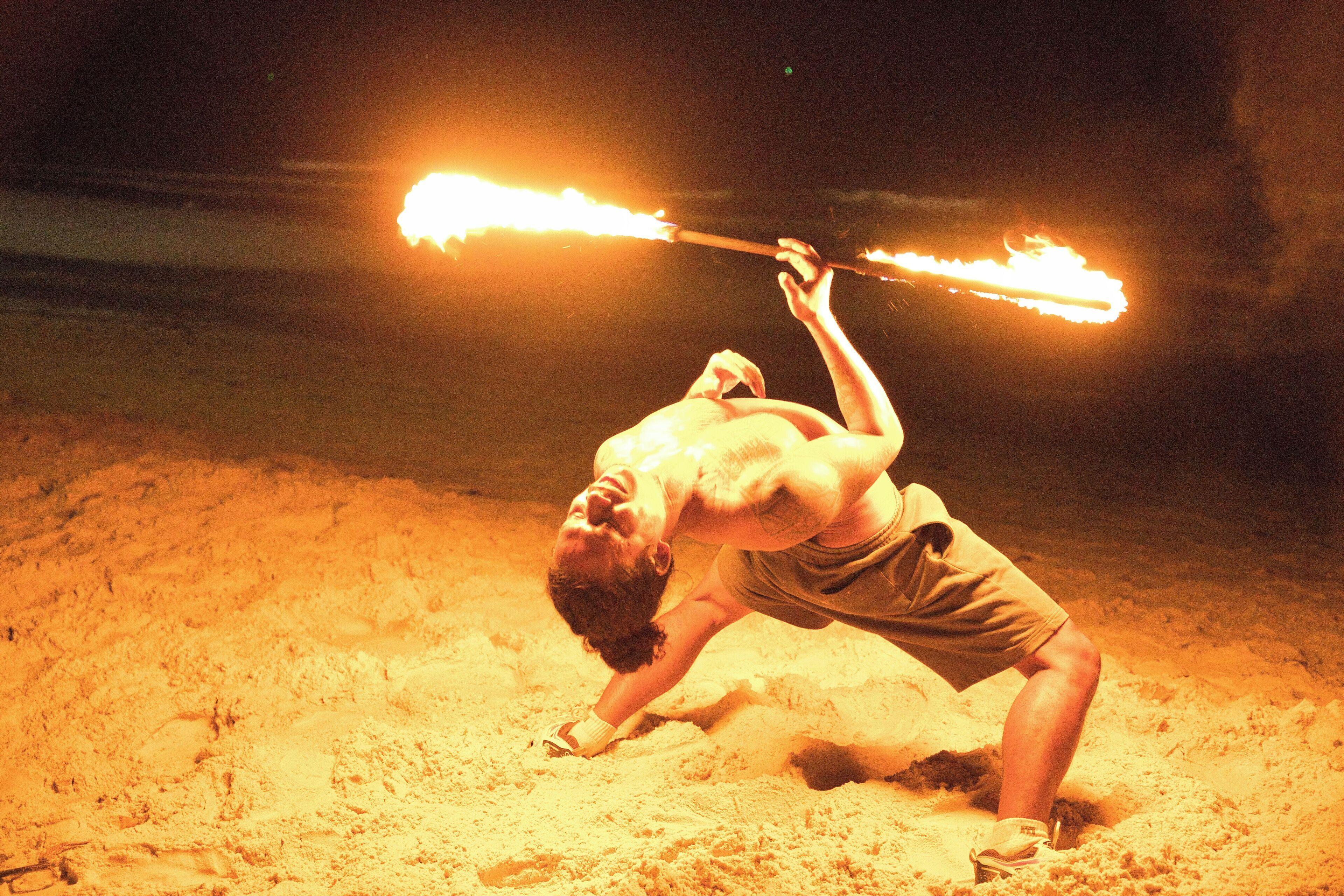 A person performing a fire-breathing act with a flaming baton on a sandy surface at night.