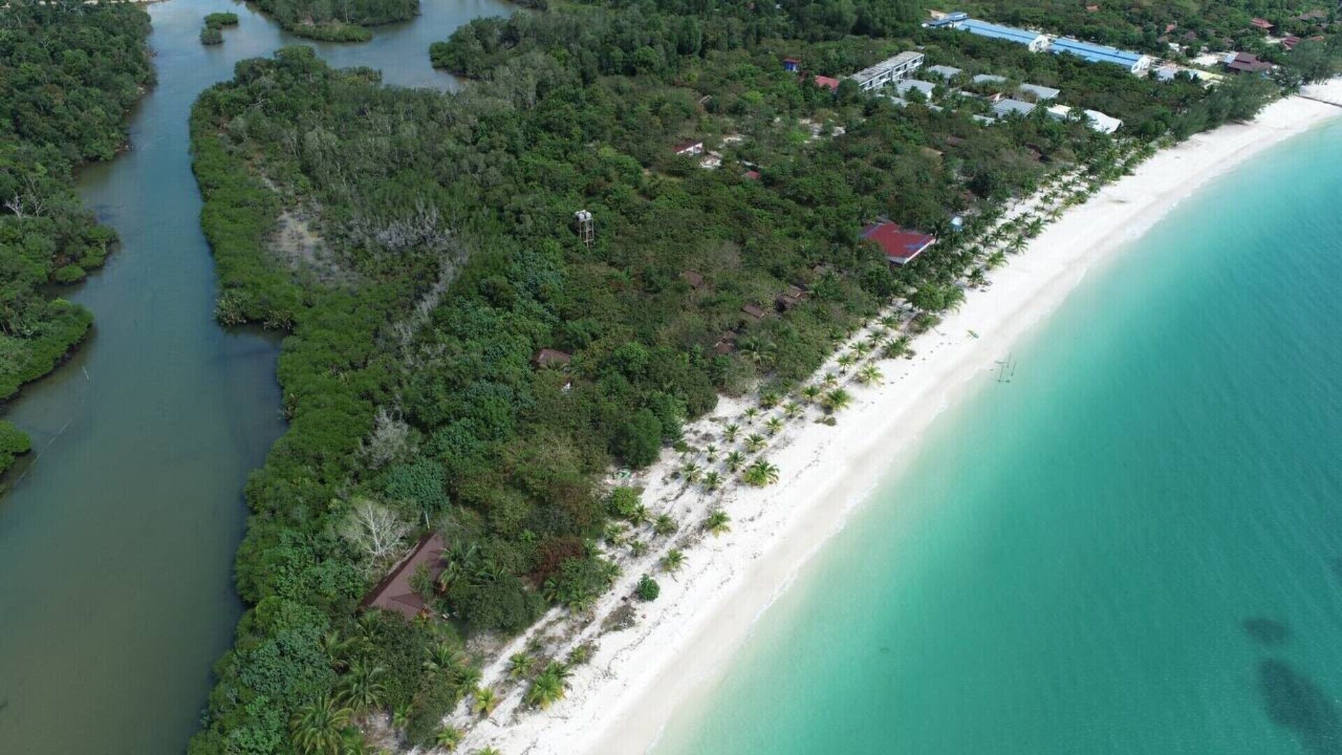 An aerial view of a coastal area with a beach, dense vegetation, and scattered buildings.