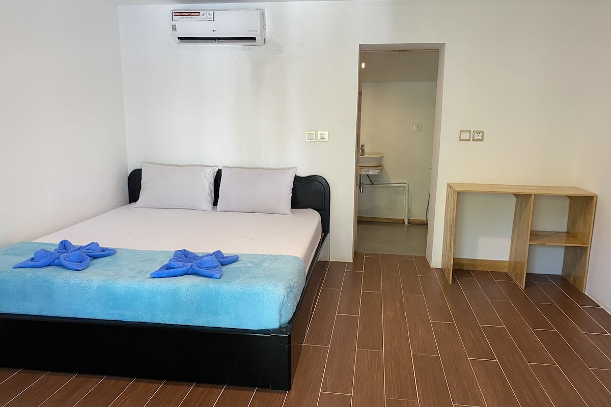 A hotel room with a bed, an air conditioning unit, a wooden floor, and a small wooden table.