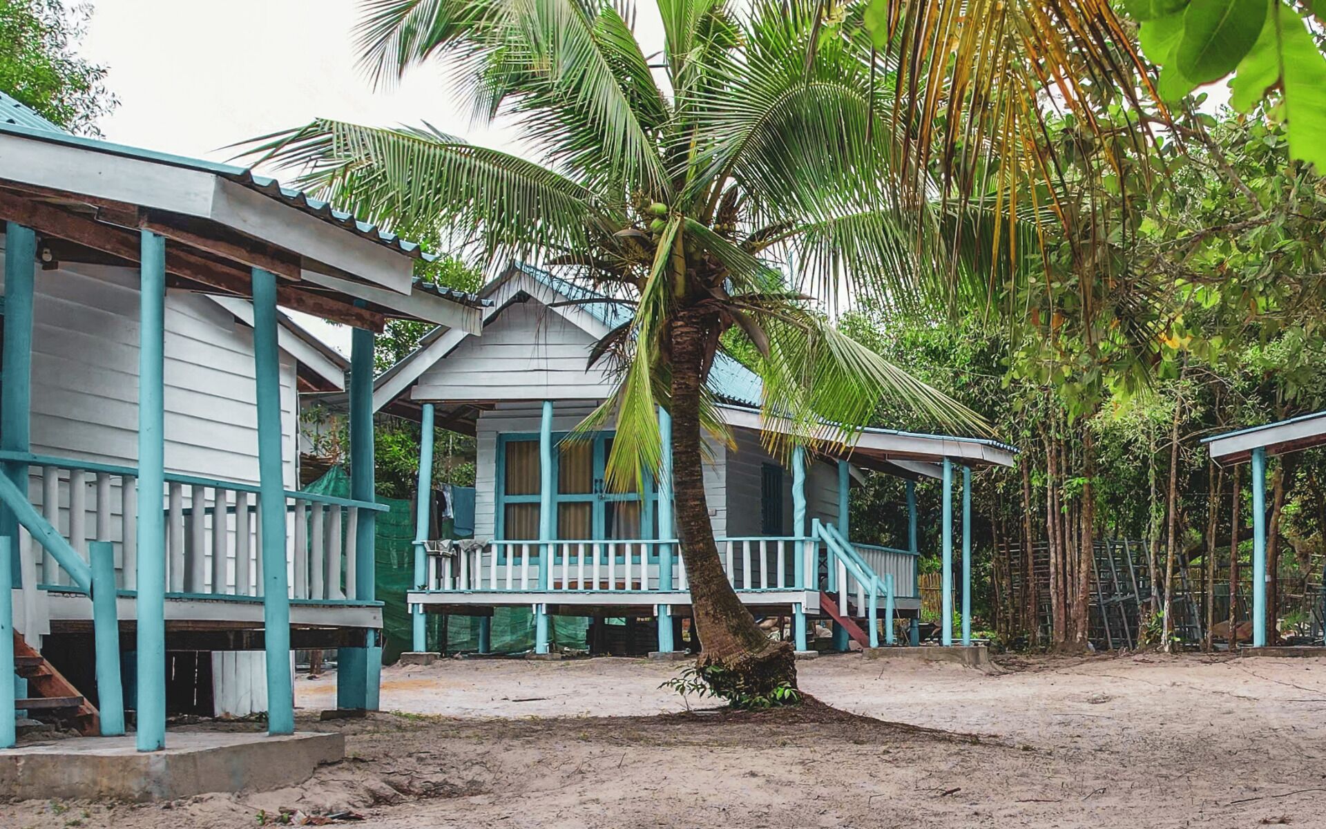 A bungalow with a porch, surrounded by palm trees and a dirt ground.