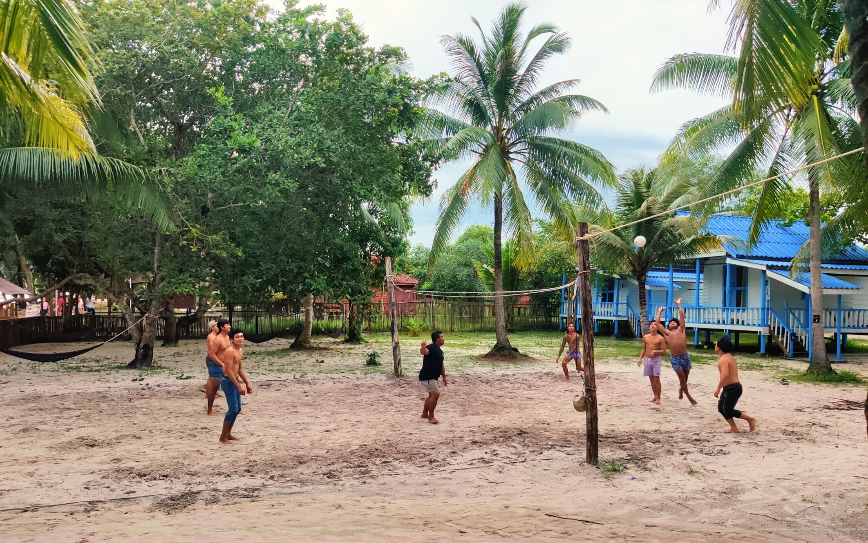 Cancha de voleibol de arena rodeada de palmeras y un edificio azul con un porche.