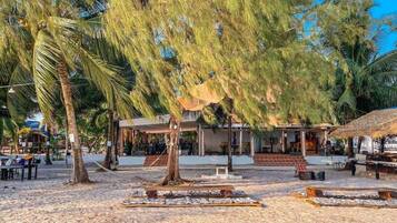 A beachside area with a covered seating area, palm trees, and a building with a thatched roof.