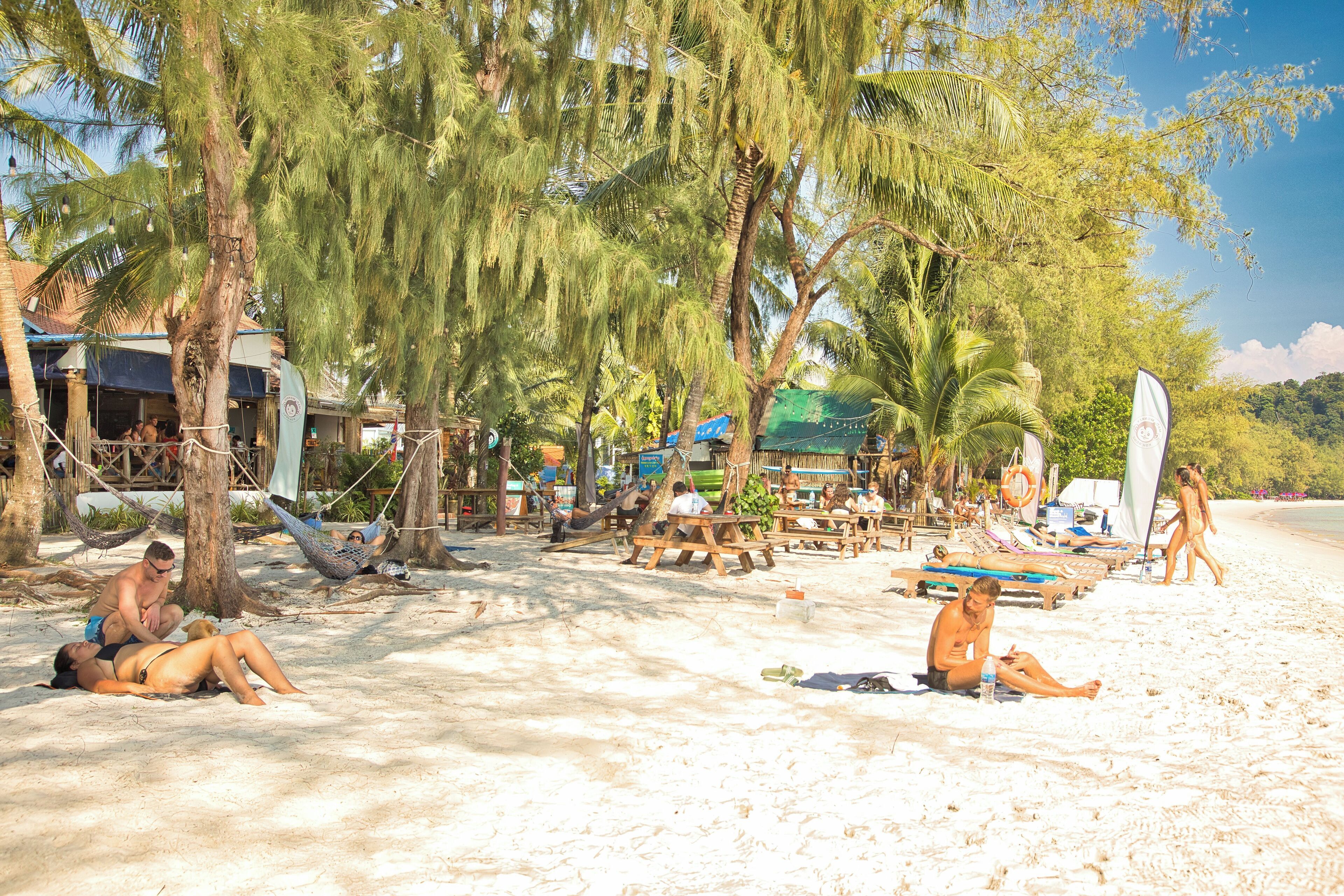 Uma cena de praia com pessoas relaxando na areia, redes e cadeiras de praia.