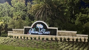 Un panneau indiquant « Palm Coast » et arborant un logo représentant un palmier, le tout entouré de verdure et d’un mur de pierres.