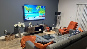 Un salon moderne avec un canapé gris, un fauteuil orange et une table basse. Un téléviseur à écran plat est fixé au mur et diffuse une image d’une plage tropicale.
