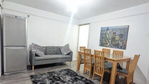 Ein kompakter Wohnraum mit grauem Sofa, Holzesstisch und Stühlen, einem schwarzen, flauschigen Teppich und einem Stadtbild an der Wand.