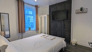 Ein Schlafzimmer mit Bett, einem fensterbelaubten Fenster, einem flachbildfernseher an einer dunklen Wandverkleidung und einer kleinen Pflanze auf einem Regal.