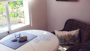 Een kamer met een ronde tafel, een rieten stoel, een kussen met een bloemenprint, een glas water en een raam met uitzicht op groen.