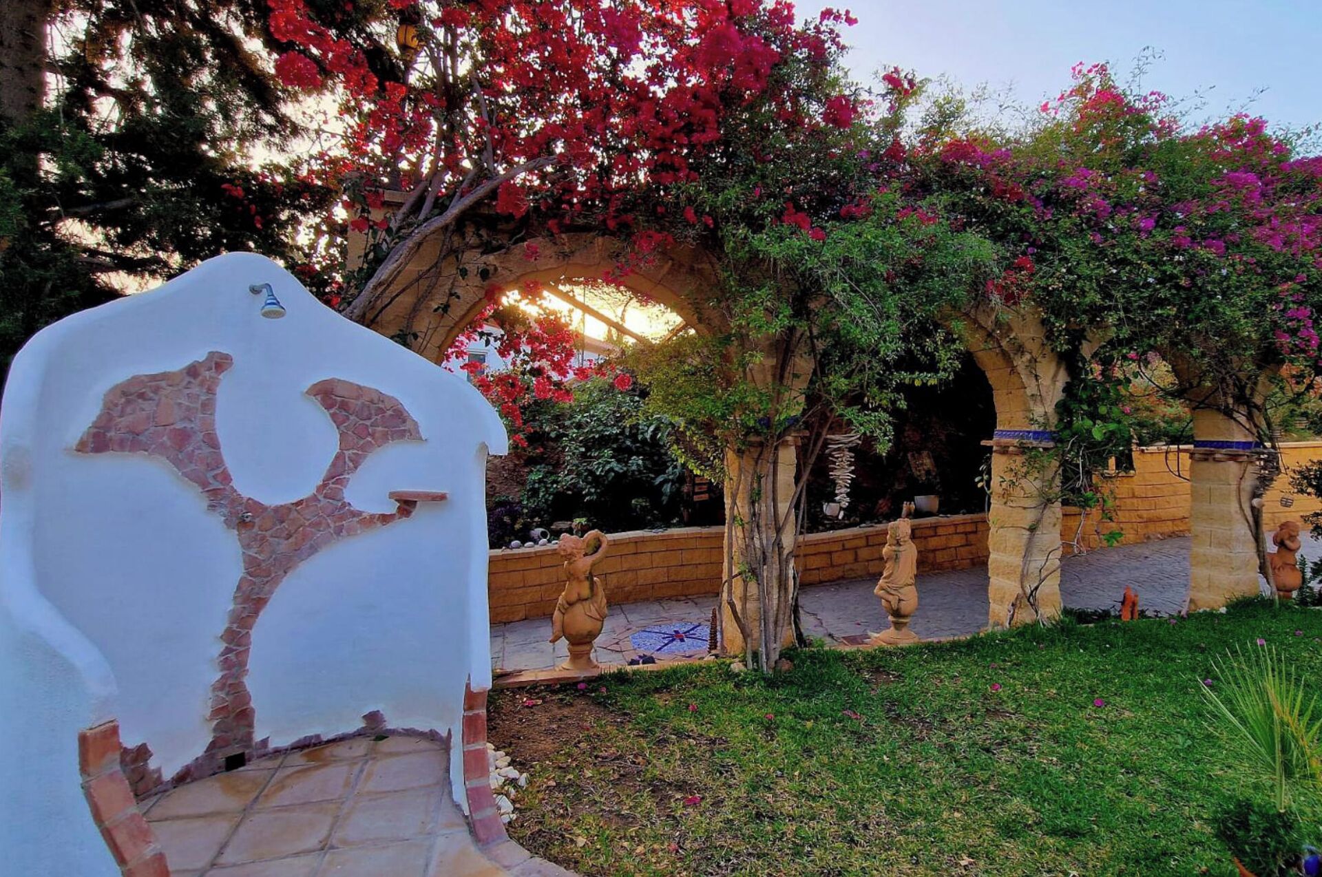 Un jardín con un muro blanco que presenta un relieve de piedra, una pérgola de madera y vistosas flores de buganvilla.