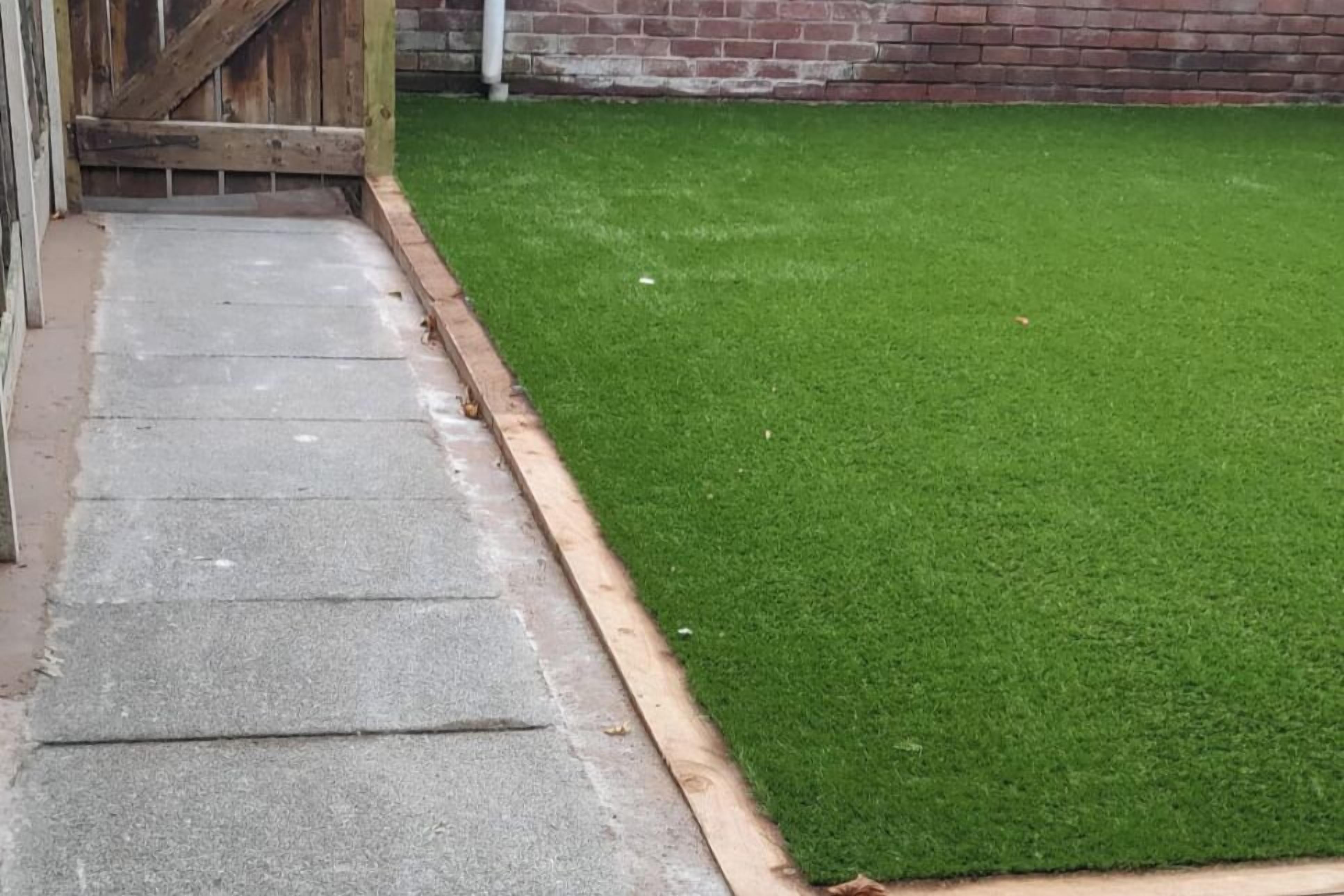 Uma trilha de concreto leva a um gramado verde, delimitado por uma cerca de madeira e uma parede de tijolos.