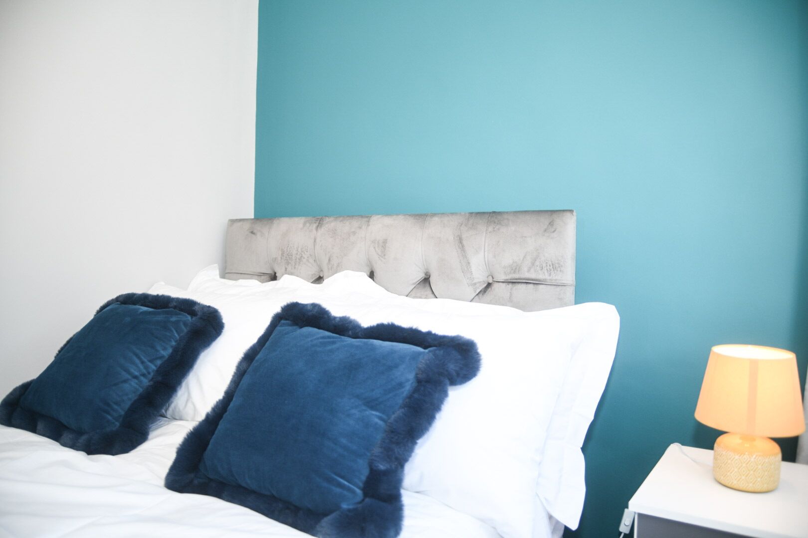 A bed with white bedding and two blue pillows, a grey tufted headboard, and a bedside lamp on a white nightstand.