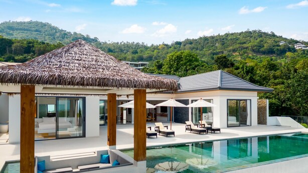 A modern resort with a pool, thatched-roof structure, and a view of lush hills.