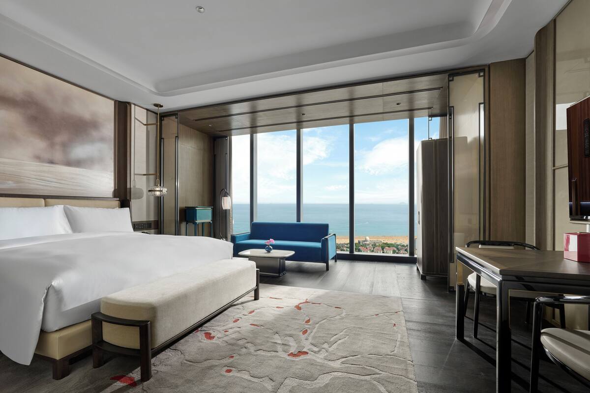 Habitación de hotel moderna con una cama grande, zona de estar con vistas al mar y una mesa de comedor.