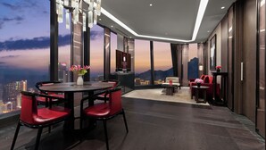 Habitación de hotel moderna con vista panorámica de la ciudad, una mesa de comedor redonda con sillas rojas y una araña.