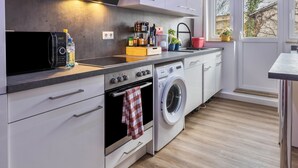 A modern kitchen with a microwave, oven, washing machine, and a window with a view of buildings.