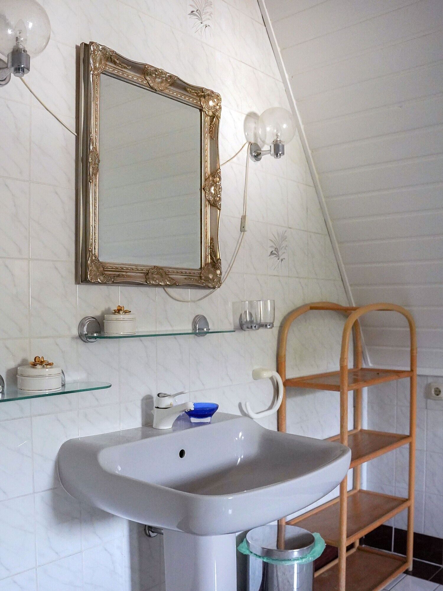 Un baño con un lavamanos blanco, un espejo con marco dorado y un estante de madera.
