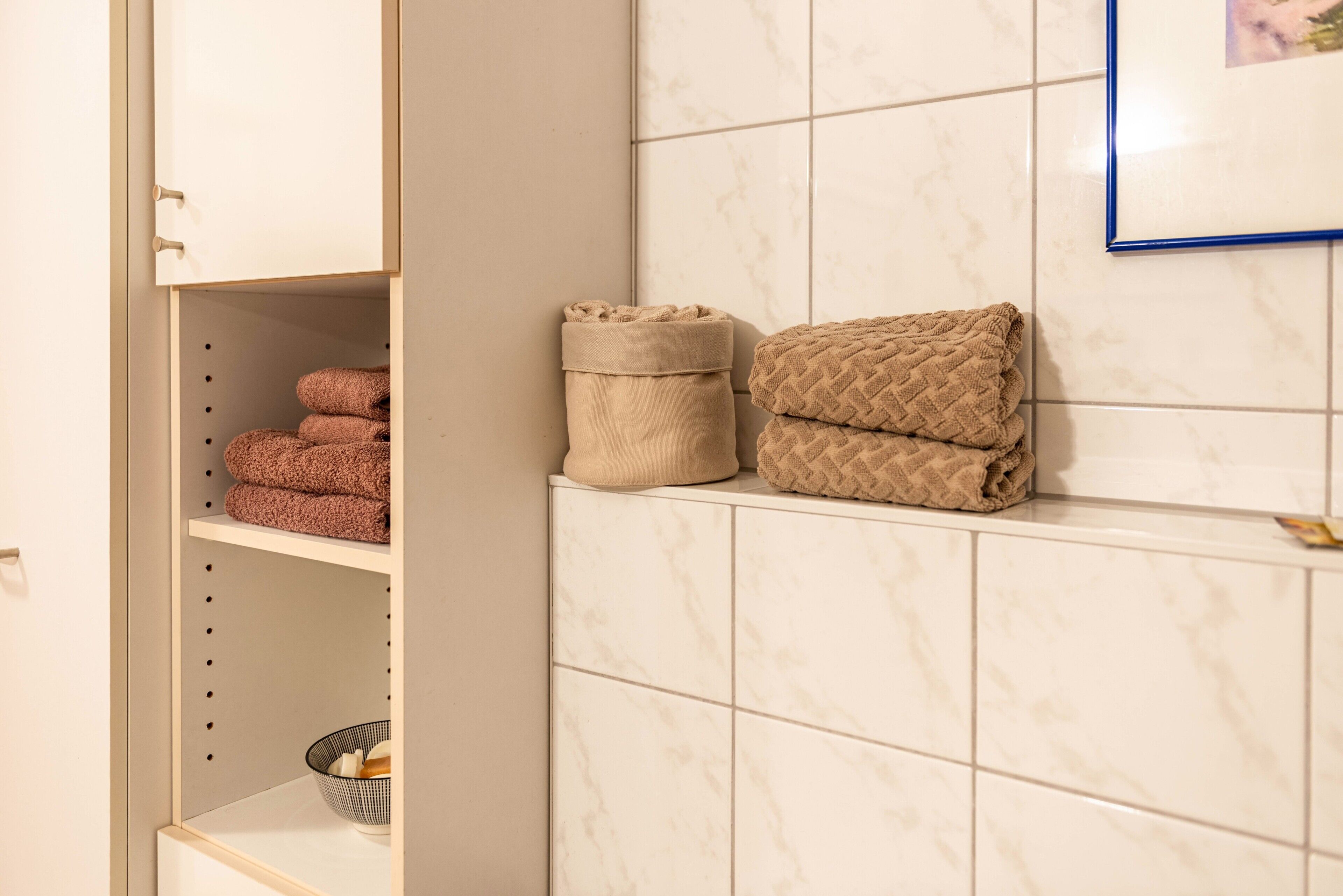 Ein Badezimmer mit weiß gefliesten Wänden, einem Handtuchhalter mit gefalteten Handtüchern und einem Ablagefach mit einer Schüssel.