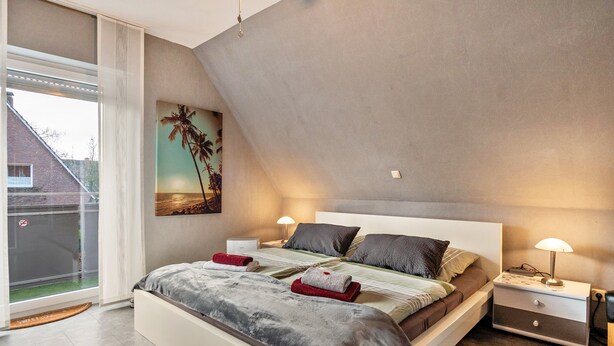 Une chambre à coucher avec un lit, des tables de chevet, un tableau et une vue sur l’extérieur, visible à travers une grande fenêtre.