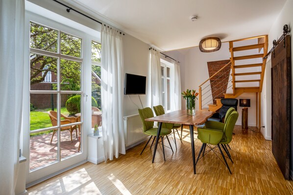 A dining area with a wooden table and green chairs, a staircase, and a view of the garden.