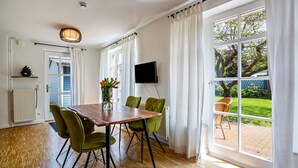 A dining area with a wooden table and green chairs, a TV mounted on the wall, and a large window with curtains. - Holiday Home 'Altes Fischerhaus Döse' with Private Garden and Wi-Fi (Cuxhaven)