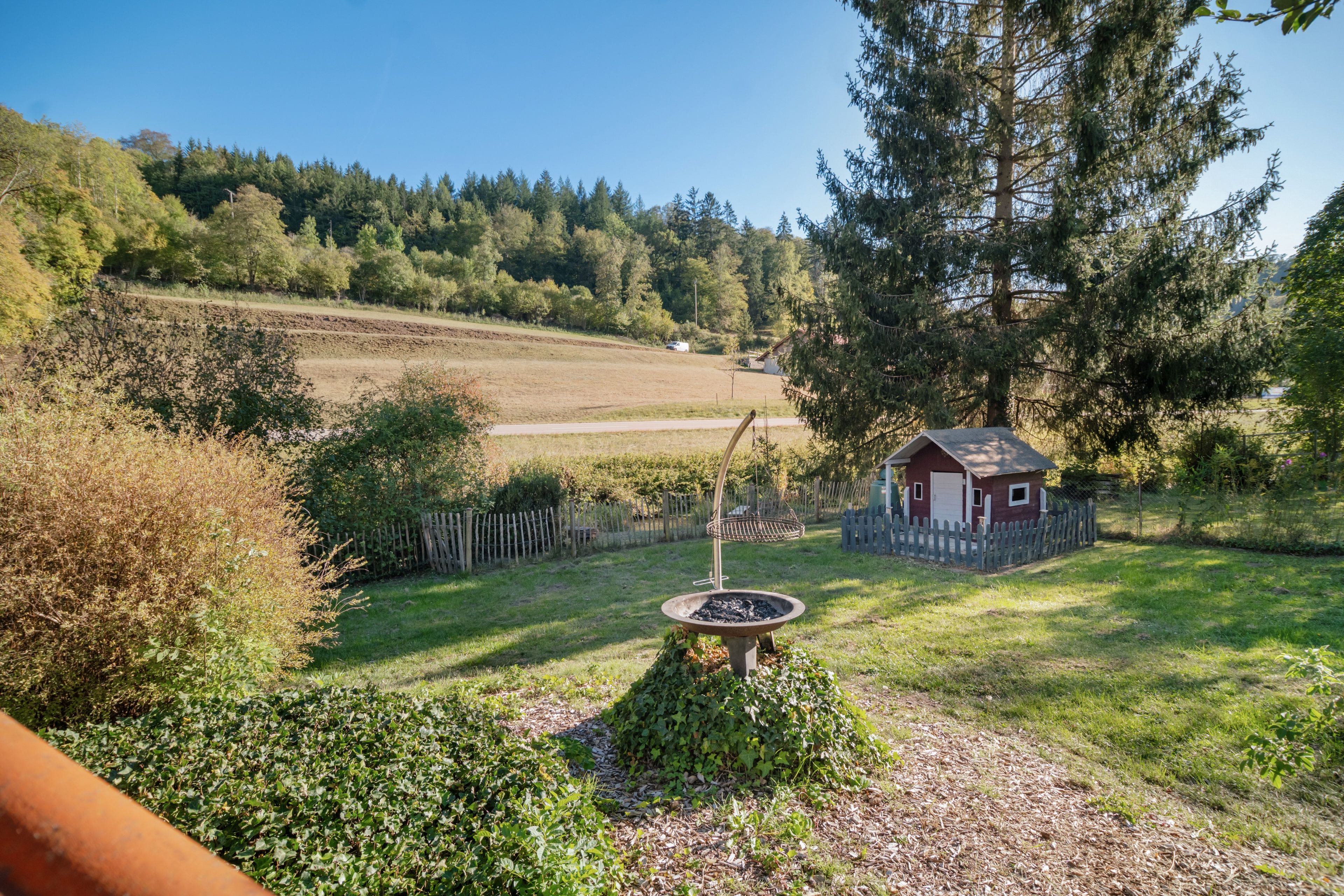 Un paysage rural avec une petite construction en bois, une mangeoire à oiseaux en métal et une zone délimitée par une clôture.